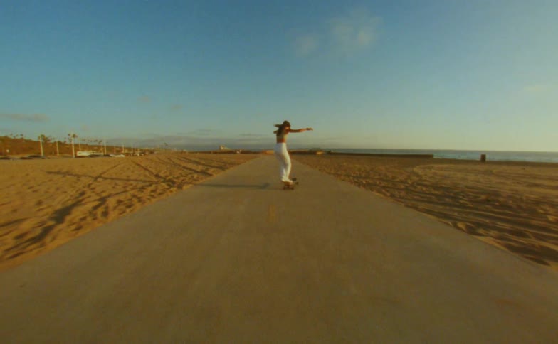 A teenage girl raises her arms in the air as she rides her skateboard on a road near the beach