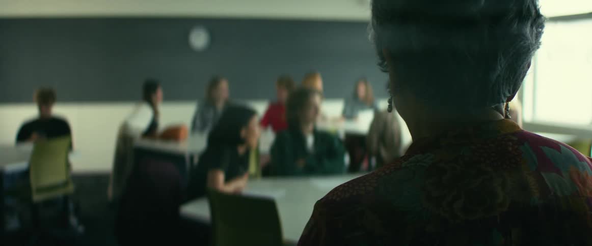 A teacher looks over her students as they talk leisurely at tables in a classroom during the day