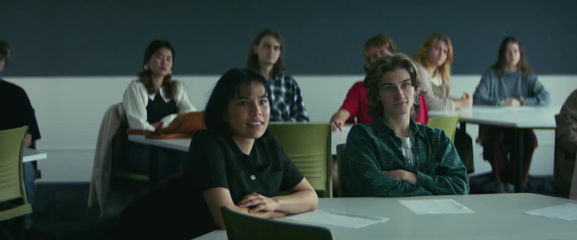 Most students sitting at tables in classroom seem bored but one smiles