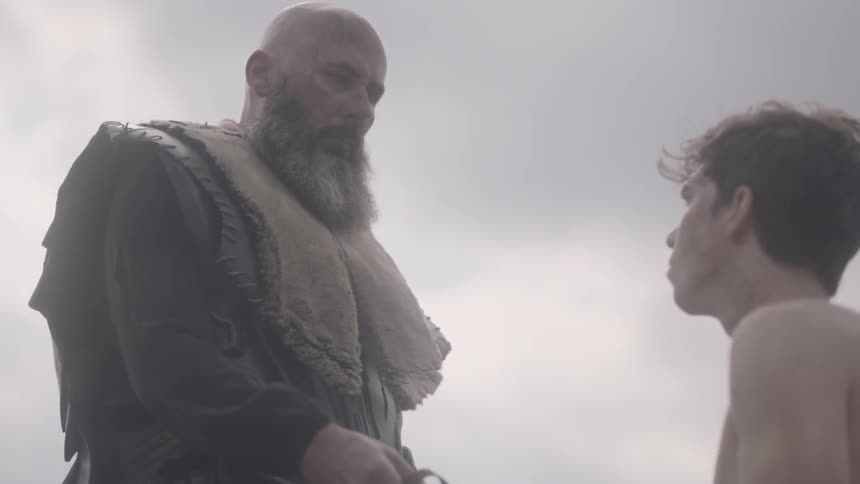 A bald man walks over to another man and hands him a sword
