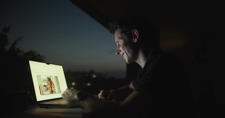 A man is sitting at a table with his laptop talking on video chat with a woman