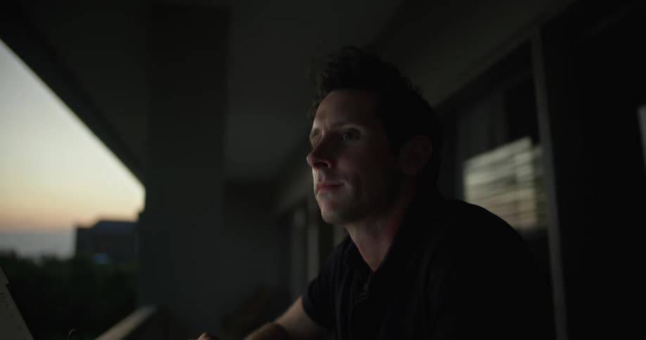 A man sits outside at a table typing on a laptop and looking around seemingly thinking