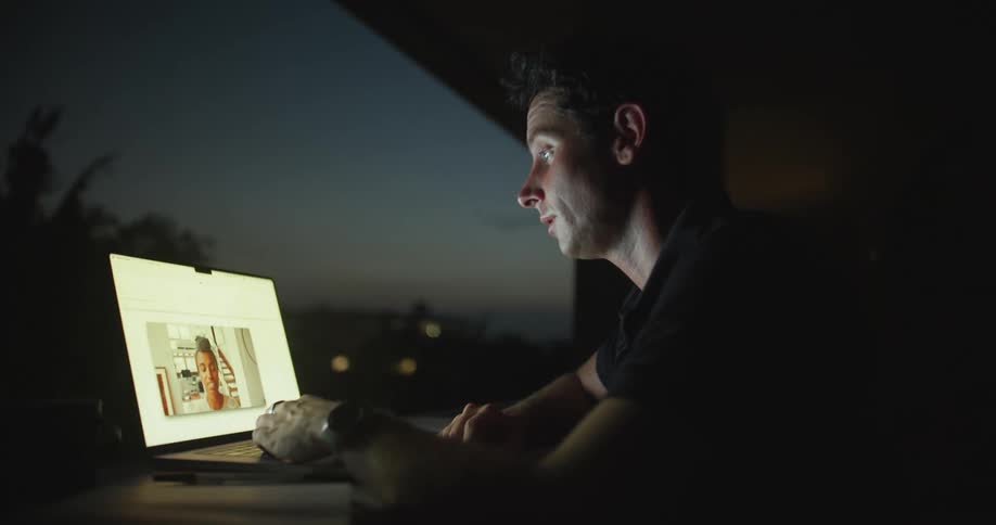 Man sitting at the desk on balcony having a video call with a woman on his laptop during night