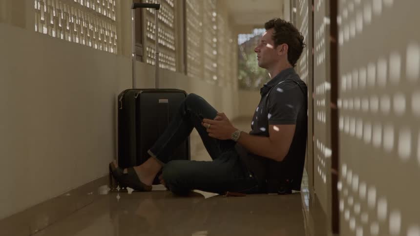 Man sitting on the floor in the hallway next to his suitcase using his cellphone