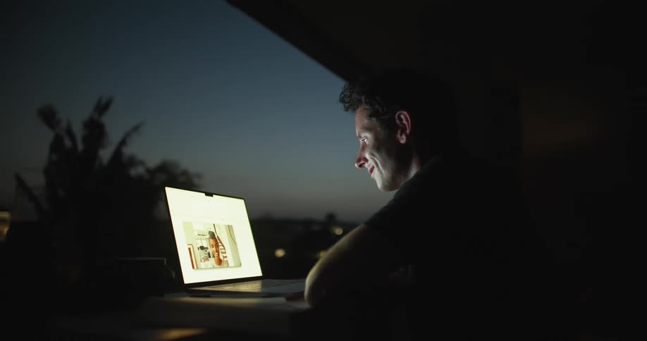 Man sitting at the desk in his balcony during the night having a video call with a woman on his laptop