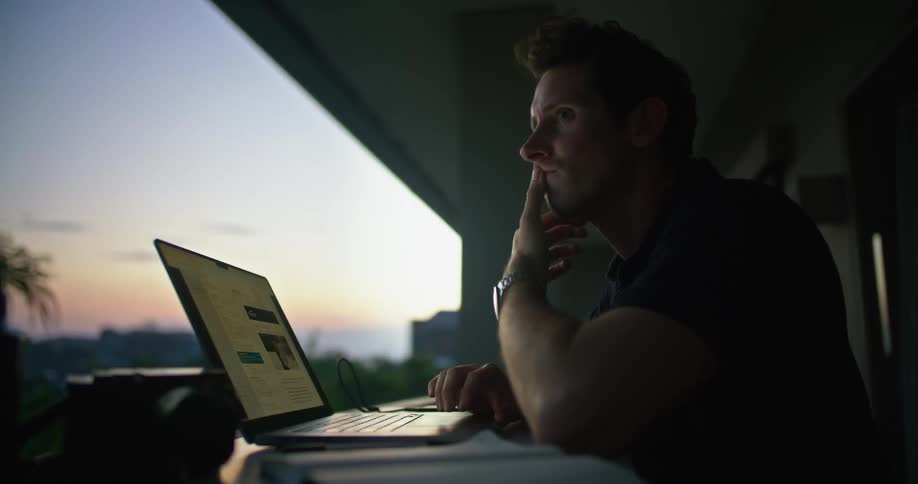 Thoughtful man sitting at the desk in his apartment balcony using his laptop at dusk