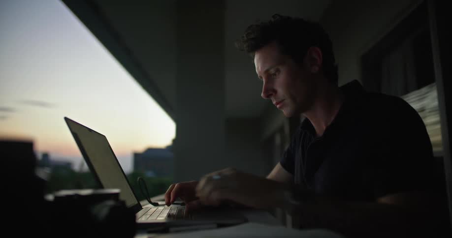 Man sitting on a dark room near a balcony at home working on a laptop at evening