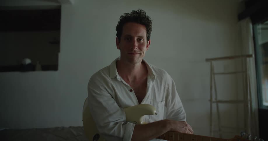 A man is sitting down with his arm resting on his electric guitar and he turns his head and smiles