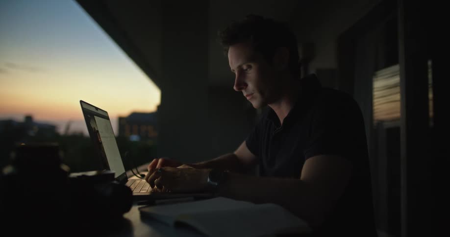 A man is sitting at a table in front of a window looking at a book as he types on a laptop computer