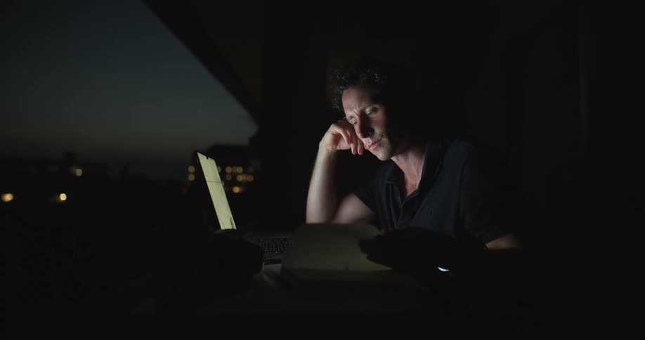 A man is sitting at a table in front of a window looking at a book as he types on a laptop computer and he rubs his face with his hand