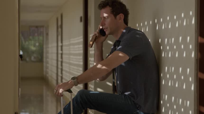 Man leaning against the hallway wall with his suitcase by his side talking on the phone