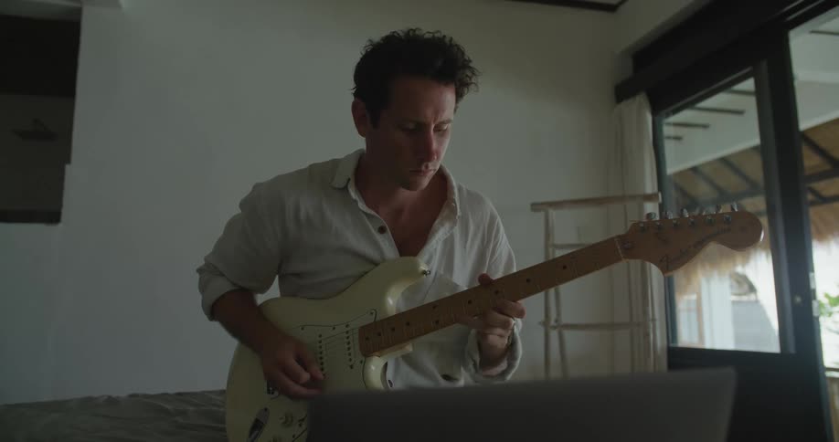 Man with white shirt sits on bed playing white guitar looking at laptop in bedroom