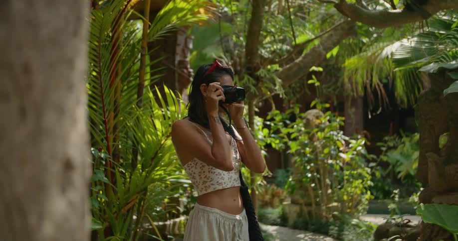 A woman is standing in a rain forest and as she smiles as she lifts her camera to her face and takes a picture