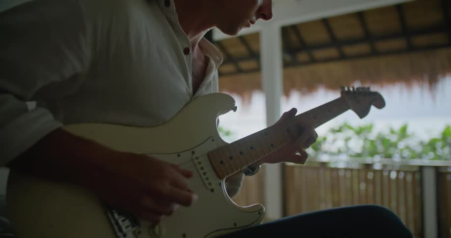 Man sitting on the bed in his room playing a guitar next to the balcony