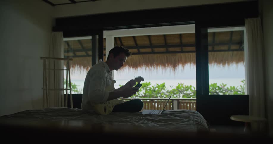 A man sits on a bed on front of large open windows playing on a guitar and looking at his phone