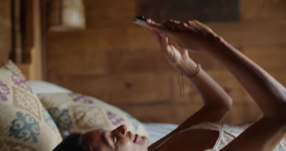 Young happy woman resting in bed browses her smartphone in the bedroom at home