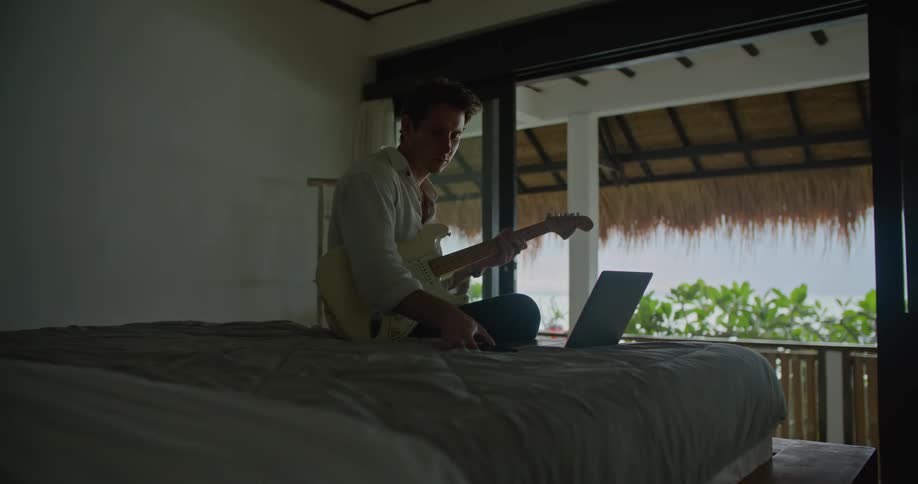 Man sitting on bed in his bedroom playing guitar and using his laptop and smartphone