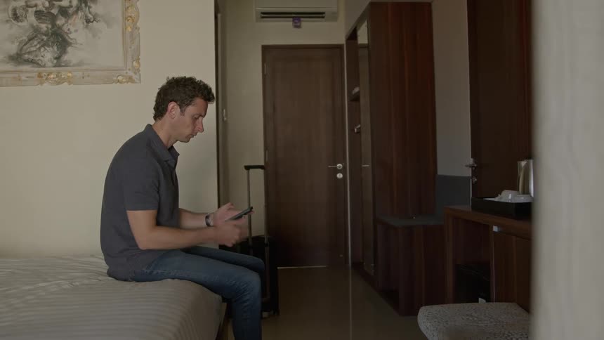 Young man sitting on the bed of a hotel room waiting for a call on his smartphone