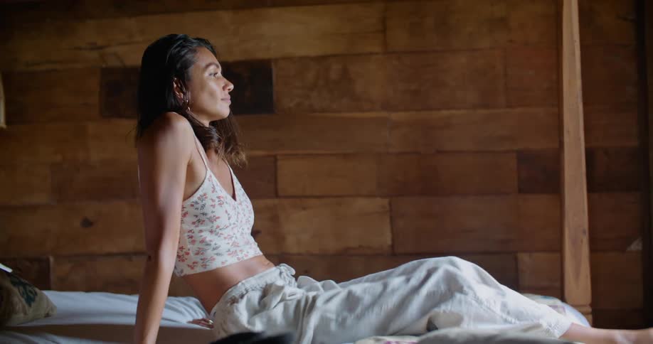 Sunlight shines upon a woman sitting on a bed as she leans back with and looks out the window