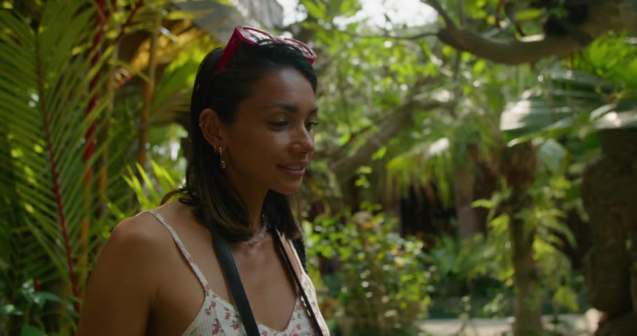 A woman is standing in a rain forest and as she smiles she lifts her camera to her face and takes a picture and then walks away