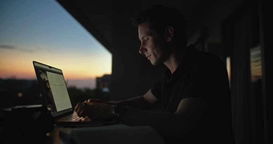 Man sitting at the desk on the balcony using his laptop while looking through some papers during sunset