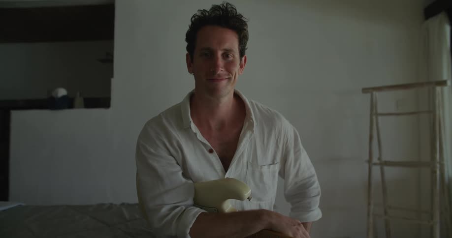 Man with white shirt and white electric guitar sits on bed smiling