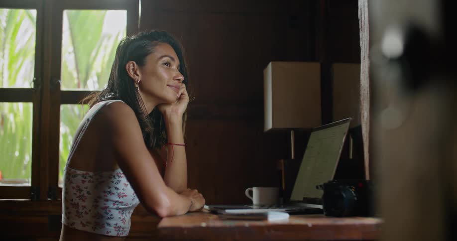 Young happy woman sitting at a desk at home thinking and typing on a laptop