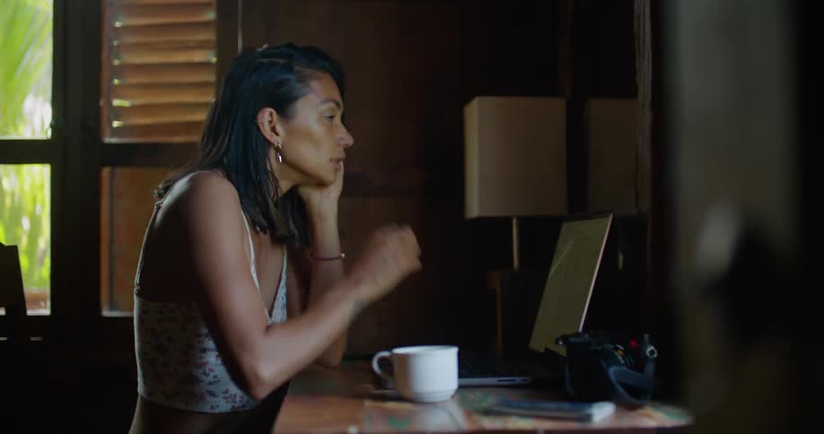 Woman with straight hair earrings printed halter blouse and tattoo types on laptop thinking