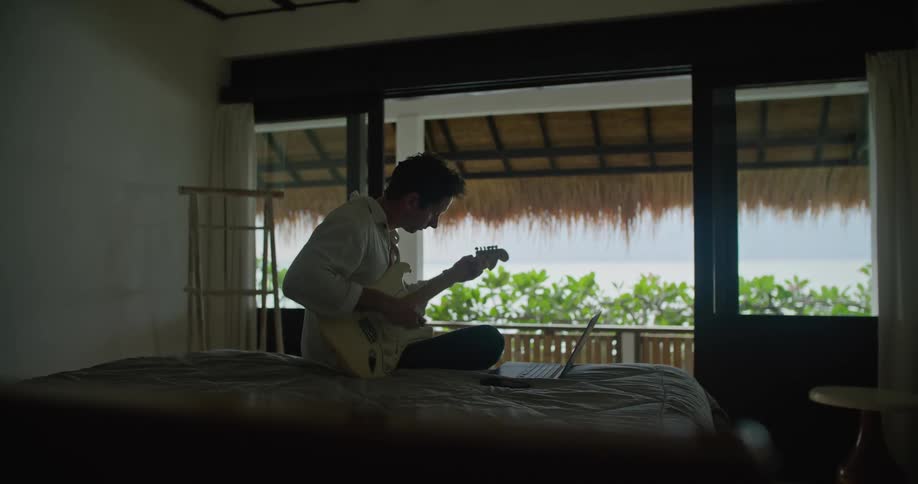 A man sitting on a bed playing guitar with his laptop open and tropical breeze blowing