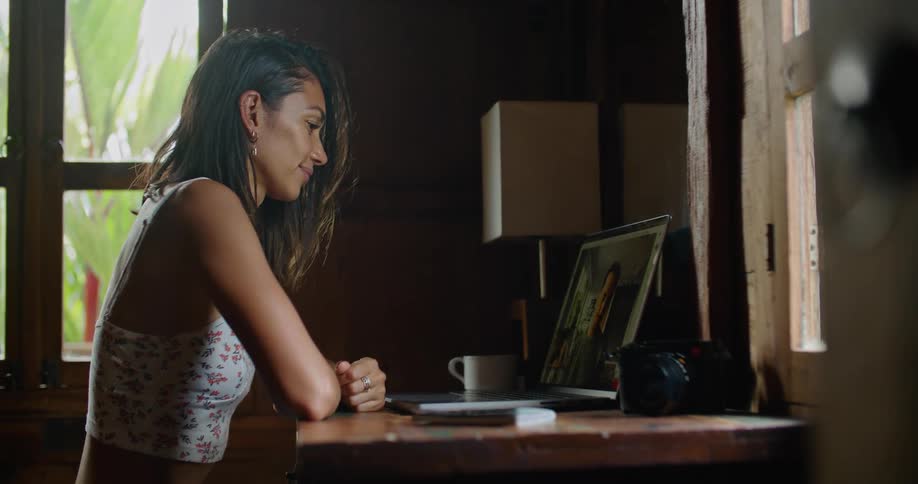 Young woman sitting on a desk at home talking to someone on a video call through a laptop