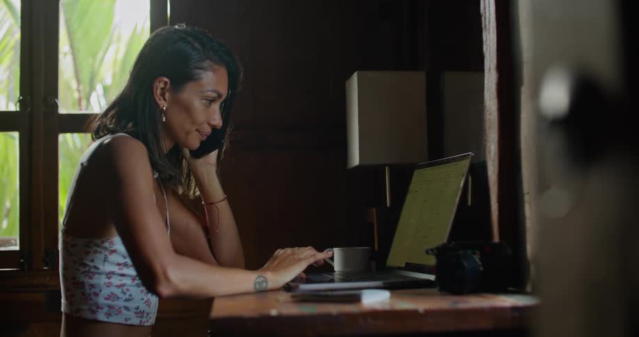 A woman is sitting at a table with a laptop computer as she talks on the phone and types on the keyboard