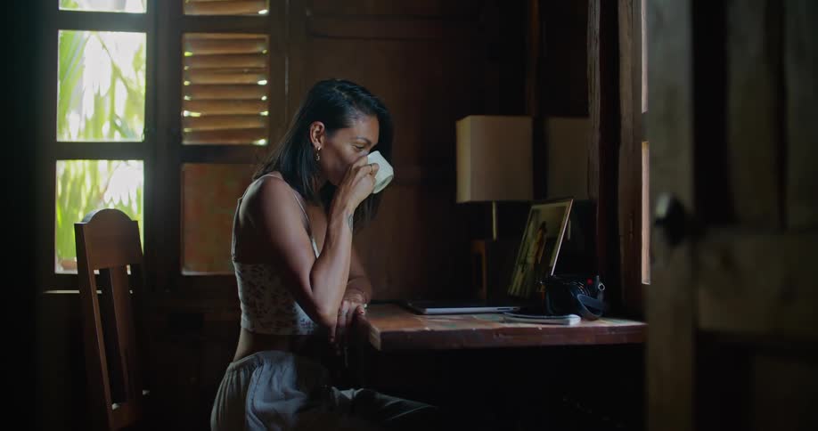 A woman is sitting down in front of a laptop computer drinking coffee and having a video conversation and waving to that person