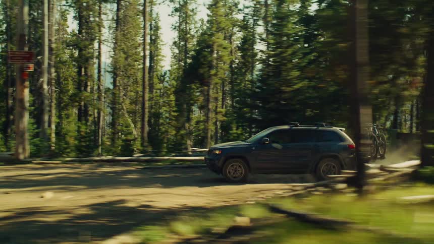 Car drives along dirt path in conifer forest kicking up dust