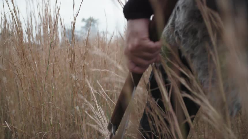 A man walks through tall grass while carrying a large axe