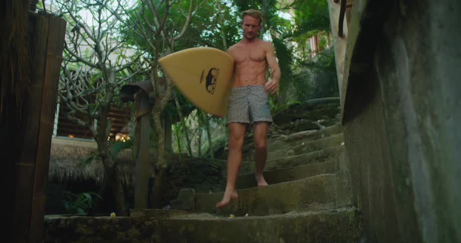 A man walks barefoot down stone steps carrying his surfboard with abs on display