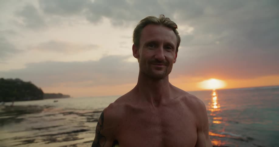 A man is standing on a beach smiling as waves roll in and the sun sets in the distance