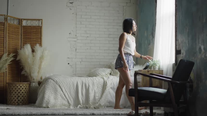 A woman walks across her chic bedroom puts down her phone and looks out the window