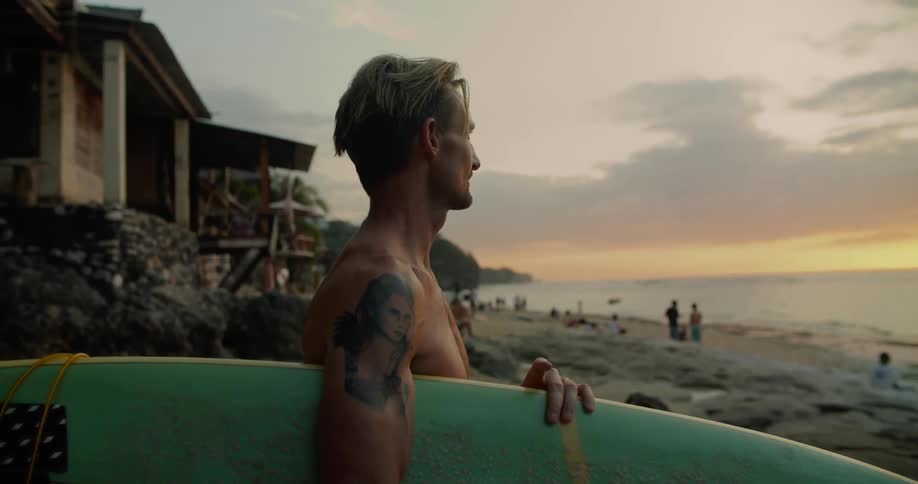A man standing on the beach holding his surfboard tapping his fingers as he looks at the water