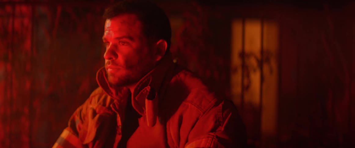 A firefighter with soot on his face takes off his helmet looking dejected in the red light glow