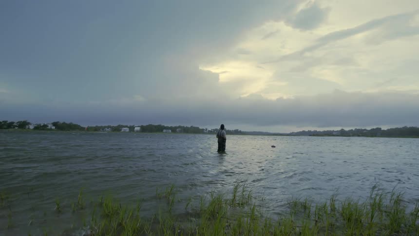 A man wearing overalls is standing in a lake as the water moves by him and he looks in the distance under a cloudy sky