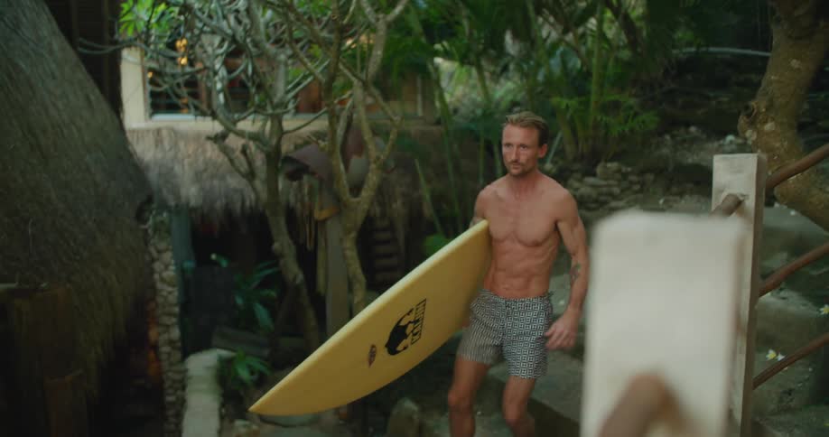 A man carrying his surfboard walks barefoot down stone steps toward the beach