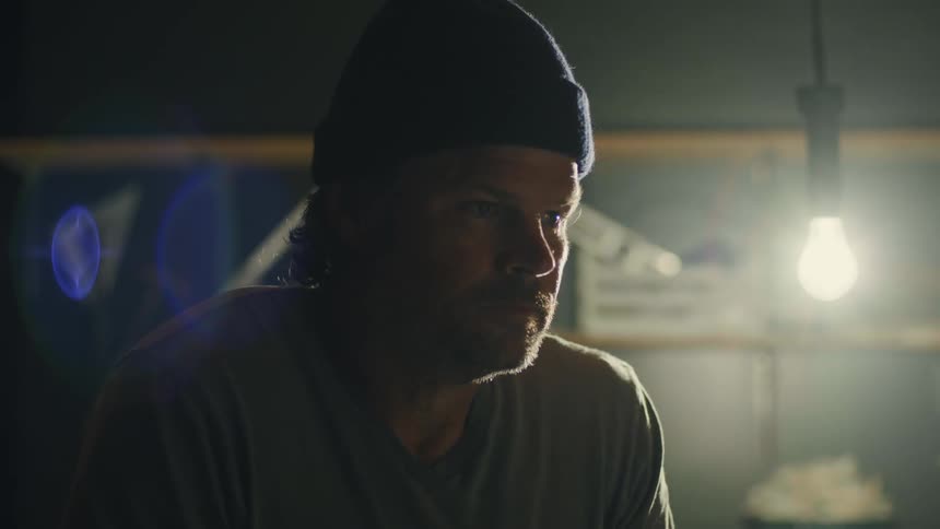Man wearing knit hat works in dim room lit by an electric bulb