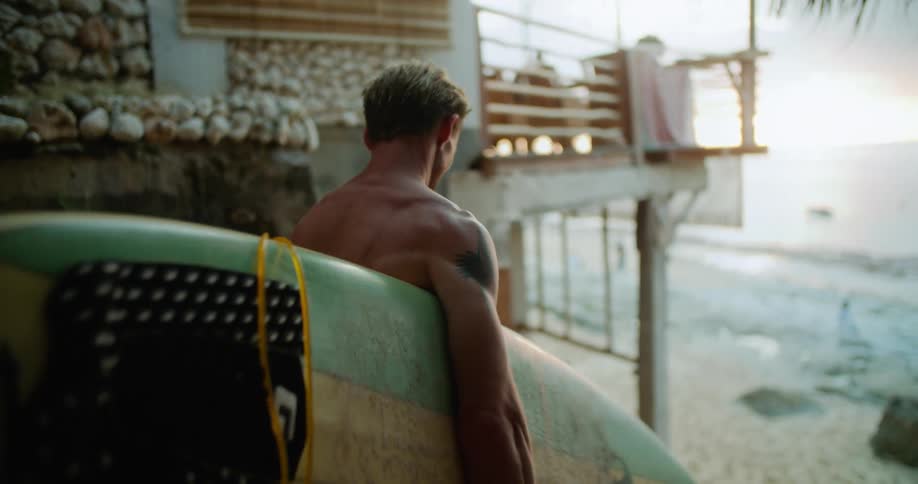 A man is standing next to a building holding his surfboard and looking at the beach then he walks down toward the water