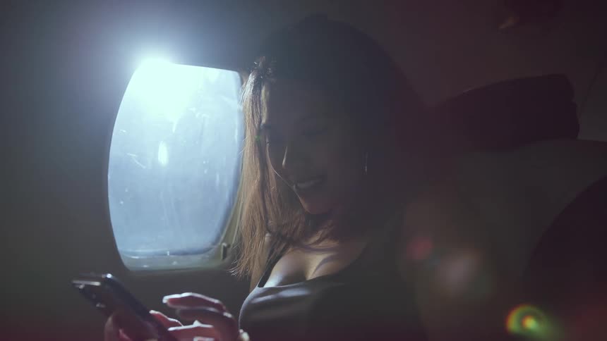 A woman is sitting in an airplane seat next to a window and texting on her phone with a smile on her face