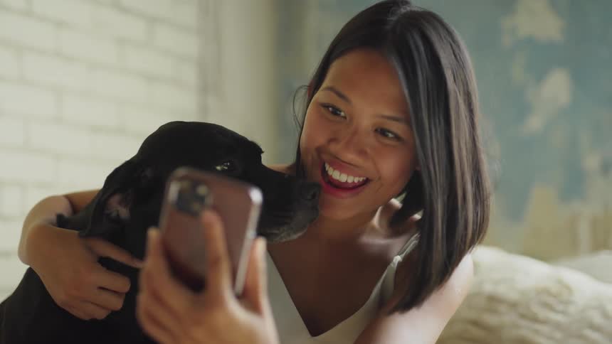 A woman sitting with her arm around her dog pets him and then takes a selfie with her cell phone and waves at the phone