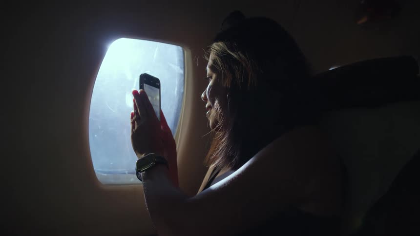 Woman sitting near window in airplane takes photograph out the window using her smartphone