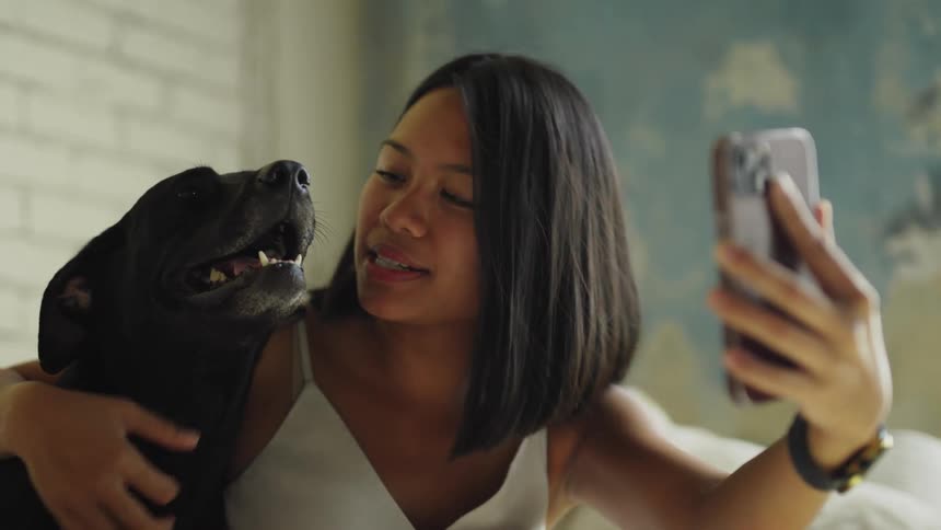 A woman smiles and talks on a video call on her phone while hugging and petting her black lab