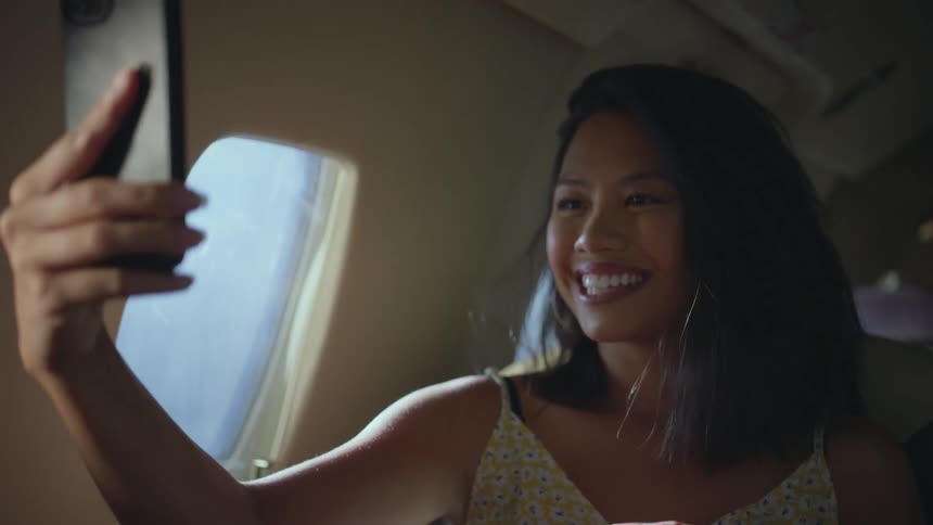 A woman is smiling and taking selfies with her cell phone while flying on an airplane