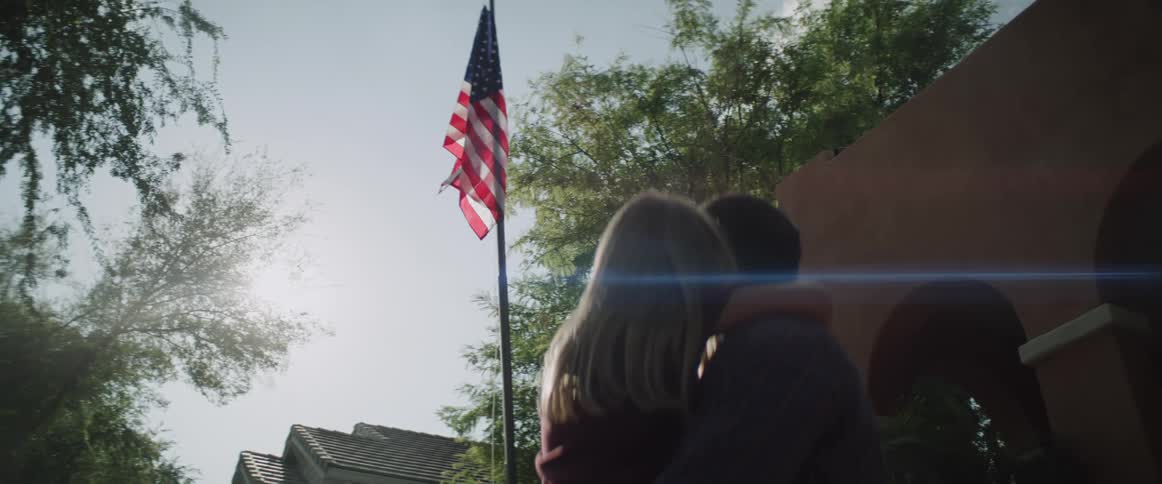 A man holds his daughter as they watch an american flag