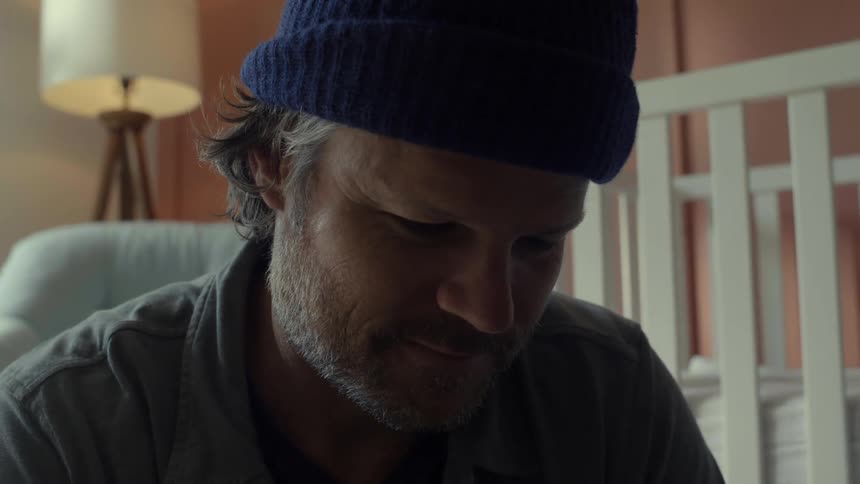 Mature bearded man wearing a blue beanie sitting near a crib on a dimly lit bedroom